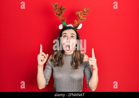 Une jeune fille hispanique portant un chapeau de noël à cerf a été stupéfaite et a surpris de regarder vers le haut et de pointer avec les doigts et les bras relevés. Banque D'Images