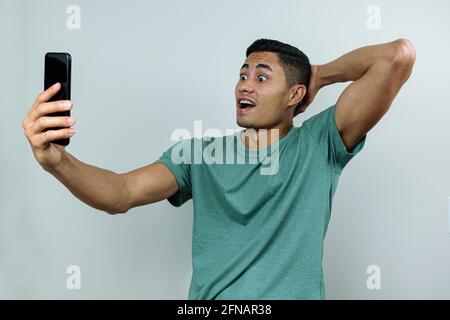 Jeune homme brésilien, tenant et regardant le smartphone, avec les yeux larges et l'autre main sur le dos du cou (surpris). Banque D'Images