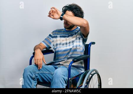 Beau homme hispanique assis sur un fauteuil roulant couvrant les yeux avec le bras, regardant sérieux et triste. sans vue, cache et rejet concept Banque D'Images