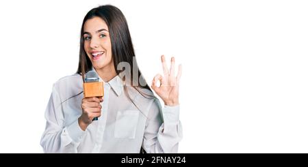 Jeune brune adolescente tenant le microphone journaliste faisant ok signe avec les doigts, souriant sympathique gestante excellent symbole Banque D'Images