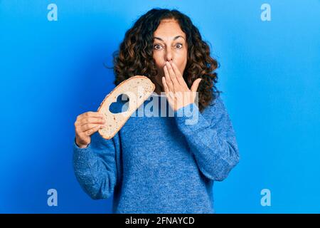 Femme hispanique d'âge moyen tenant le pain de forme de coeur couvrant la bouche avec la main, choqué et peur de l'erreur. Expression surprise Banque D'Images