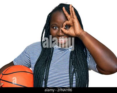 Jeune femme noire avec des tresses tenant le ballon de basket-ball souriant heureux panneau ok avec la main sur l'œil en regardant à travers les doigts Banque D'Images