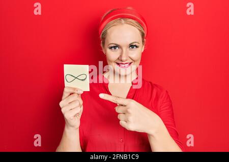 Jeune femme blonde tenant le symbole infini rappel sourire heureux pointage avec la main et le doigt Banque D'Images