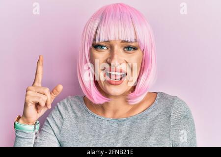 Jeune femme caucasienne portant une perruque rose pointant du doigt vers le haut avec une idée réussie. Sortie et heureuse. Numéro un. Banque D'Images