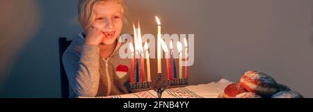 Fille éclairant des bougies sur la menorah pour les vacances traditionnelles d'hiver juive Hanukkah. Enfant célébrant le festival des lumières de Chanukah. Beignets Sufganiyot Banque D'Images