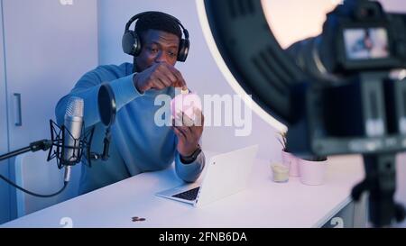 Homme portant un casque et tenant une banque de pigeons expliquant quelque chose devant la caméra. Mettre des pièces de monnaie dans la tirelire. Un microphone et un ordinateur portable sont placés sur la table. Enregistrement vidéo. Photo de haute qualité Banque D'Images