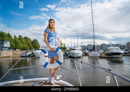 Jeune femme heureuse d'apparence caucasienne dans une robe à rayures bleues debout sur un yacht posant avec une bouée de sauvetage dans sa main, sur le fond d'un ciel bleu avec des nuages par une journée ensoleillée d'été Banque D'Images