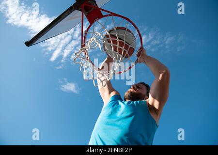 Joueur de basket-ball. Sports et basket-ball. Un jeune homme saute et jette une balle dans le panier. Ciel bleu et cour en arrière-plan. Banque D'Images