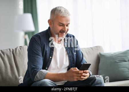 Homme mûr souriant aux cheveux gris assis sur un canapé, à l'aide d'un téléphone portable Banque D'Images
