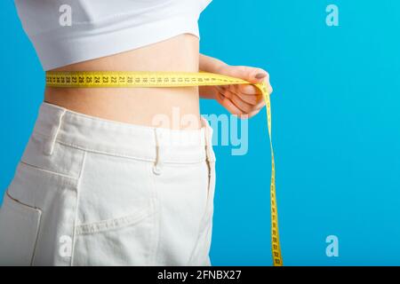 Une femme près du corps mesure sa taille à l'aide d'un ruban de mesure. Concept de perte de poids en forme de corps sain. Taille fine petit ventre dans un grand pantalon en denim blanc Banque D'Images