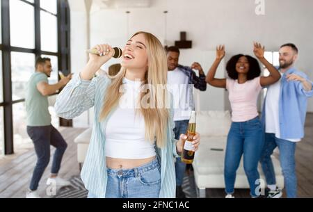 Jolie jeune femme blonde chantant le karaoké à la maison, tenant une bouteille de bière, dansant et s'amusant avec un groupe d'amis Banque D'Images