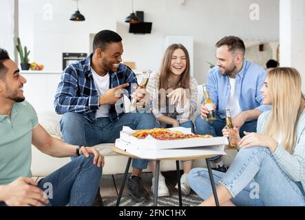 Groupe de divers amis masculins et féminins mangeant de la pizza à la maison, buvant de la bière, ayant la conversation sur la fête étudiante Banque D'Images