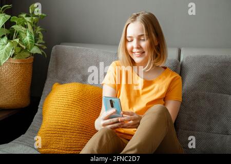 Femme souriante utilisant un smartphone à la maison. Ado blonde fille hippster recherche chat sur le téléphone dans la chambre sur le canapé. Une jeune femme utilise l'application mobile pour discuter en ligne Banque D'Images