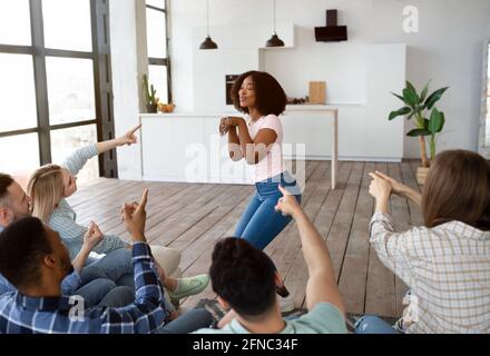 Groupe diversifié de jeunes meilleurs amis jouant des charades dans la salle de séjour, essayant de deviner le mot sur la partie étudiante Banque D'Images