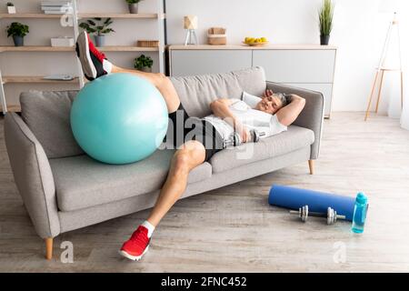 Fatigue homme mûr reposant sur un canapé après un entraînement dur actif. Sportif fatigué avec haltères et ballon de fitness sur le canapé Banque D'Images