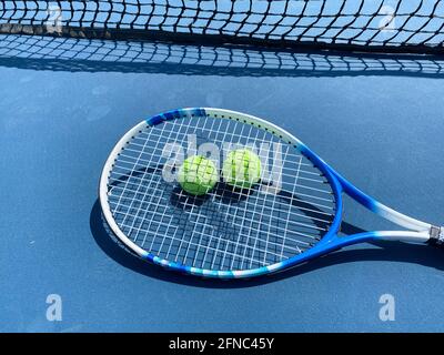 Une raquette de tennis située au-dessus de deux balles de tennis jaunes sur un court bleu près du filet de tennis. Banque D'Images