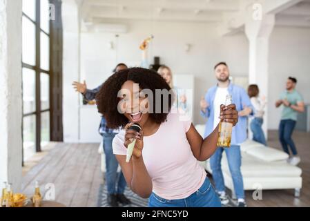 Jeune femme noire avec bouteille de bière et microphone chantant karaoké avec d'autres étudiants à la maison Banque D'Images