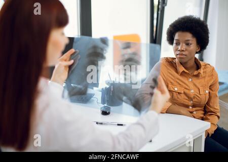 Vue de l'épaule du médecin, tenant l'examen thoracique par rayons X, expliquant les résultats au patient. Concentrez-vous sur la jeune femme africaine à l'écoute des recommandations du médecin Banque D'Images