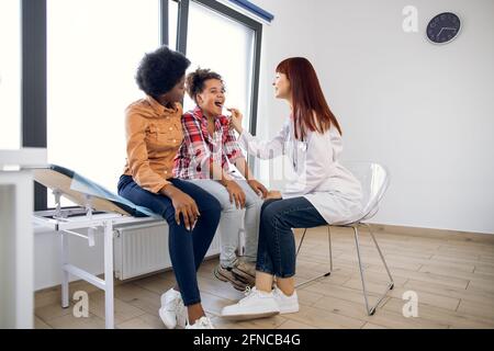 Prise de vue en longueur d'une jeune femme laryngologiste médecin examinant la gorge d'un enfant afro-américain avec un dépresseur de langue. Jolie jeune mère noire soutenant sa fille au bureau du médecin Banque D'Images