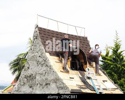 Deux couvreurs couvrant le toit d'une maison en argile décorative dans le parc parmi les arbres verts du sud contre un ciel nuageux Banque D'Images