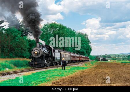 Strasburg, PA, USA - 15 mai 2021 : un homme Amish regarde une locomotive à vapeur de la route ferroviaire de Strasburg se prépare à s'éloigner d'une plantation de pique-nique dans r Banque D'Images