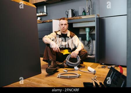 Jeune plombier en unifrom assis sur le sol par ouvert armoire lors de la réparation de l'équipement Banque D'Images