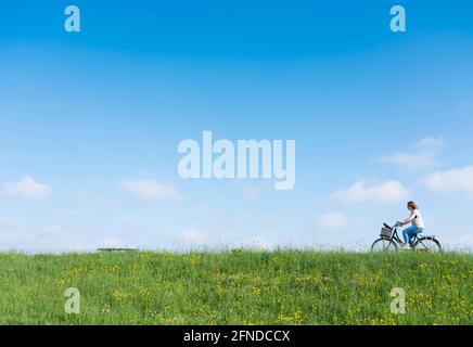 une femme fait du vélo sur une digue herbeuse avec des fleurs sous le bleu ciel Banque D'Images