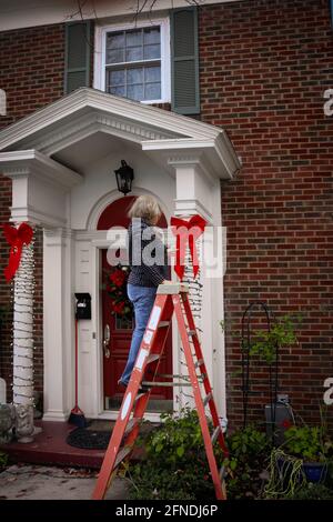 Femme en échelle redresse les lumières et un ruban de décorations de Noël De pilier de porche sur maison traditionnelle Banque D'Images