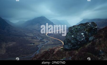 Paysage matinal avec feux de voiture sur la route de montagne menant à Glencoe et un grand rocher en premier plan. A82 route en face de Buachaville Etive Mor avant l'aube. Écosse Banque D'Images
