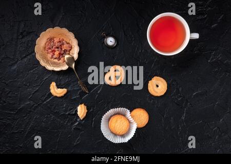 Biscuits au beurre danois avec une tasse de thé et une montre montrant le temps de thé, au-dessus plat couché sur un fond noir Banque D'Images