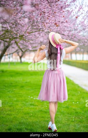 Belle femme en chapeau de paille marchant dans un jardin d'été avec des cerisiers en fleurs. Fille portant une jupe longue rose Banque D'Images