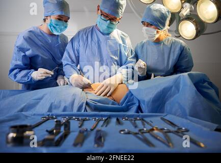 Groupe de médecins portant des gants stériles et des masques protecteurs faisant de la chirurgie plastique en salle d'opération. Équipe chirurgicale effectuant une chirurgie esthétique alors que le patient est allongé sur la table d'opération près des outils médicaux. Banque D'Images