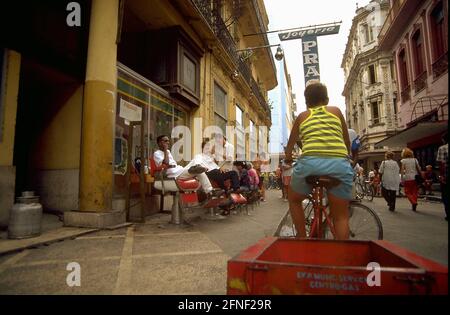 Un cycliste avec une remorque s'approche d'un salon de coiffure qui sert des clients dans la rue à des températures tropicales. [traduction automatique] Banque D'Images