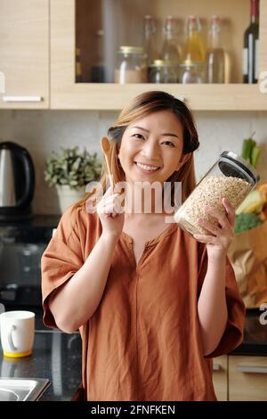 Portrait d'une jeune femme heureuse et excitée avec un pot de flocons d'avoine et une cuillère en bois dans la cuisine Banque D'Images