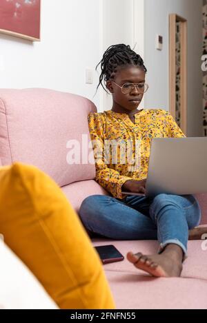 Femme indépendante afro-américaine concentrée assise sur un canapé à la maison et de taper sur netbook tout en travaillant sur le projet Banque D'Images