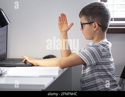 Un élève de l'école élémentaire avec des lunettes est assis à une table avec un ordinateur portable, apprend à distance, lève sa main dans une leçon en ligne. Banque D'Images