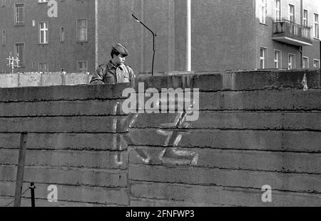 Berlin-City / GDR / 20.3.1980 Bernauer Strasse in Wedding. Les anciennes pierres murales sont remplacées par des éléments en forme de L en béton lisse. Le jeune garde-frontière est là pour surveiller les ouvriers de la construction. Pour les empêcher de fuir vers l'Occident. Du côté ouest, quelqu'un a écrit -KZ- sur le mur. Derrière le mur se trouve Berlin-Mitte. // RDA / mur / districts / Stasi / Histoire / communisme [traduction automatique] Banque D'Images