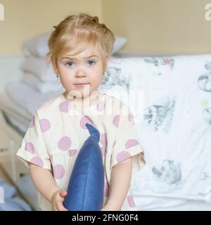 Jolie petite fille à yeux bleus dans une robe en lin à pois tenant un coussin décoratif bleu marine et regardant un appareil photo tout en passant du temps à la maison, adorable petit enfant aux cheveux dorés posant à l'intérieur Banque D'Images