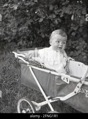 Années 1950, historique, à l'extérieur, garé sur de l'herbe, un jeune garçon assis dans une poussette de l'époque, recouverte de toile, encadrée de métal, à roues métalliques, pliante, fabriquée par Tan-Sad, Angleterre, Royaume-Uni. Fondée à Birmingham en 1922, Tan-Sad a fabriqué des jouets de construction, des chaises d'usine en métal et des fauteuils de poussée pour enfants. Dans les années 1950 et 1960, le nom Tan-Sad est devenu si bien connu qu'il est devenu un nom générique pour les poussettes. La société a plié en 1975. Banque D'Images