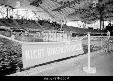 'Au bord de la piscine est accroché un signe sur une chaîne avec l'inscription 'ne pas sauter dans'. En arrière-plan, vous pouvez voir un garçon sauter dans la piscine depuis une tour de plongée. [traduction automatique]' Banque D'Images