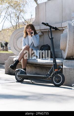 Jeune femme afro-américaine pleine de corps joyeuse en manteau bleu avoir une conversation téléphonique tout en étant assis sur un banc de pierre au printemps stationner à proximité du scooter Banque D'Images