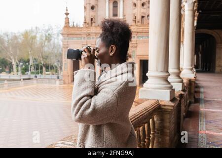 Vue de côté jeune photographe afro-américain en pelage chaud prendre des photos de bâtiments historiques de la ville avec un appareil photo moderne au début du printemps da Banque D'Images