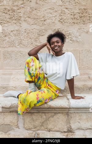 Femme afro-américaine souriante dans une tenue tendance, assise contre le mur et en regardant loin en été ensoleillé Banque D'Images