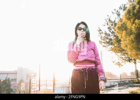 Femme millénaire en mode et lunettes de soleil fume la cigarette contre bâtiment urbain sous ciel blanc Banque D'Images