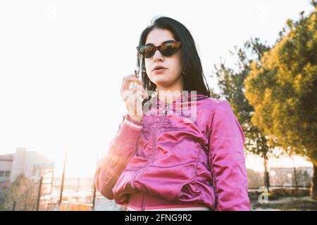 Femme millénaire en mode et lunettes de soleil fume la cigarette contre bâtiment urbain sous ciel blanc Banque D'Images