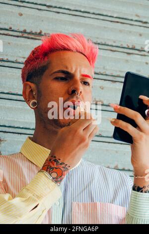 Gay élégant avec tatouage et cheveux roses appliquant le rouge à lèvres sur se retourne en regardant le téléphone portable sur fond gris Banque D'Images