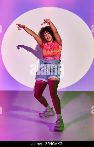 Danseuse afro-américaine pleine forme en short dansant avec les bras relevés et la languette vers l'extérieur en regardant la caméra pendant debout dans des lumières néon dans le goujon Banque D'Images
