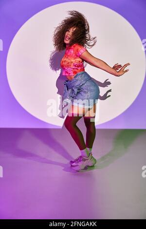 Vue latérale de la danseuse afro-américaine en short dansant en regardant la caméra avec la langue dehors tout en se tenant debout lampes au néon en studio Banque D'Images