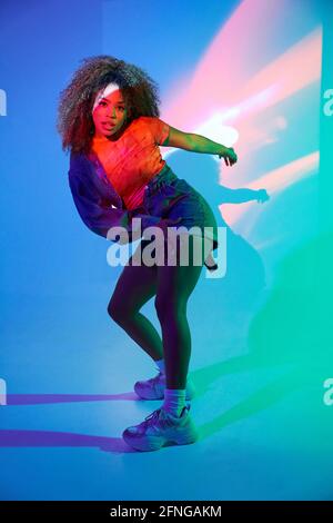 Tout le corps attrayant coupe afro-américaine femme en short dansant dans les lampes au néon en studio et regarder la caméra Banque D'Images