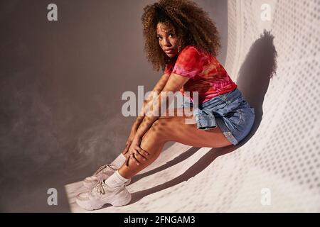 Vue latérale sur tout le corps magnifique confiante Afro-américaine femme short posé sur un mur aux courbes et regardant l'appareil photo Banque D'Images